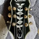 Fotografie zachycuje hlavu kytary v černé barvě s bílým lemováním. V horní části je bílý nápis Epiphone a uprostřed se nachází dekorativní vložka ve…