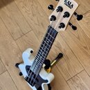 Bílá elektrická basová ukulele s dřevěným hmatníkem a hlavou. Na hlavě je černý nápis KALA UBASS. Nástroj stojí na černém stojanu s žlutým detailem a…