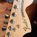 Fotografie zachycuje hlavu elektrické kytary z přírodního světlého dřeva s chromovanými ladičkami. Na dřevě je černý nápis Fender® STRATOCASTER a men…