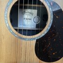 Detail akustické kytary s přírodním dřevem desky a tmavým hmatníkem. Kolem otvoru je ozdobný lem z perleti a vedle něj černý pickguard s hnědo-oranžo…