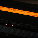 Detailní záběr na oranžový LCD displej hudebního zařízení. Na displeji je text „PATCH PLAY PR-A:012 PianoString9s center=C 4“. Pod displejem jsou vid…