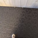Detailní záběr na černý povrch pouzdra nebo kufru s reliéfním vzorem v podobě stylizovaných spirál. Horní hrana je mírně odřená a vyjeven je kousek v…