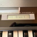 Detailní pohled na ovládací panel a klávesy syntetizátoru. Na černém glossy povrchu je bílý nápis PROGRAMMABLE POLYPHONIC SYNTHESIZER JU-1. Pod nápis…