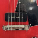 Detail červené elektrické kytary s černým pickguardem. Je vidět jeden černý humbucker s viditelnými polosnovými póly a chromovaný kobylník. Povrch pi…