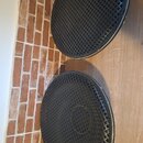Dvě černé kulaté mřížky z plastu s mřížkovým vzorem. Jsou umístěny na dřevěné podlaze vedle cihlové zdi. Na mřížkách nejsou vidět žádné značky ani ná…