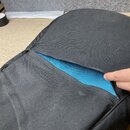 Černý textilní obal s rozepnutým zipem, který odhaluje modrou vnitřní podšívku. Povrch černého materiálu vykazuje lehké znečištění a prach. Na fotce…