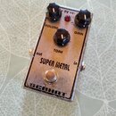 Kovový efekt pedál stříbrné barvy s nápisy SUPER METAL a BERHAT. Na horní straně jsou tři černé ovládací knoflíky s popisky VOLUME, TONE a GAIN a čer…
