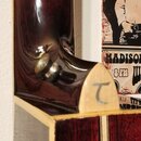 Detail krku a spoje s tělem strunného nástroje v tmavě červeném lakovaném provedení. Na světlém dřevěném prvku u spoje je viditelné stříbrné logo ve…