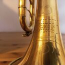 Detail mosazného zvonu trubky s viditelným opotřebením povrchu a drobnými škrábanci. Na zvonu je vyryt nápis „HULLET MUSIC CO. ST LOUIS“. Kolem textu…