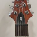 Detail hlavy elektrické kytary z hnědého dřeva s šesti stříbrnými ladičkami. Na dřevěné části je bílý nápis "PRS SE" a podpis "Paul Reed Smith". Na č…