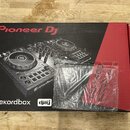 Kartonová krabice s obrázkem DJ kontroléru v černém a červeném provedení. Na krabici jsou nápisy Pioneer DJ, rekordbox a djay. Před krabicí leží průh…