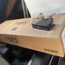 Na hnědé cardboardové krabici leží černý sustain pedál s připojeným kabelem. Na krabici je v několika místech vidět logo nord a na boční straně nápis…