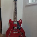 Červená polodublová elektrická kytara s tmavým hmatníkem a černým pickguardem. Na hlavě nástroje je bílý nápis Mond. Kytara má dva humbuckery, tři ov…