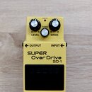 Žlutý efekt pedál s nápisy SUPER OverDrive a SD-1. Na horní části jsou tři stříbrné ovládací knoflíky s popisky LEVEL, TONE a DRIVE a červená kontrol…