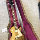 Zlatá elektrická kytara s tvarem singlecut v hnědém tvrdém pouzdře s fialovou výstuhou. Na hlavě hmatníku je bílý nápis Gibson. Kytara má dva snímače…
