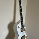 Bílá elektrická kytara s tvarem singlecut a zlatým hardwarem. Nástroj má dva humbuckery, čtyři ovládací knoflíky a bílý binding kolem hmatníku a korp…