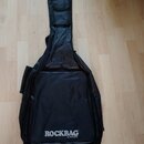 Černý poloměký pouzdro (gig bag) na basovou kytaru. V dolní části je bílý nápis ROCKBAG a pod ním menší text by Warwick. Povrch materiálu vykazuje mí…