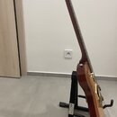 Elektrická kytara v červeně hnědé barvě s viditnou strukturou dřeva, umístěná na černém stojanu. Na noze stojanu je bílý nápis RockStand®. Povrch nás…