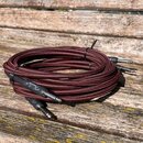 Svinutý instrumentální kabel s červeně a černě pleteným povrchem. Na jednom z černých konektorů je bílý nápis "Fender®". Kabel je stahován černým pla…
