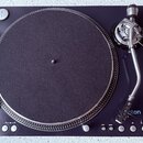Černý gramofon s kulatým talířem potaženým tmavě šedou plstí a stříbrným vlnitým okrajem. Na raménku je viditelné logo "aton" s modrým symbolem. Ovlá…