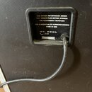 Detail zadní strany černého přístroje s textem na štítku. Je zde uveden nápis VOR ÖFFNEN NETZSTECKER ZIEHEN, PULL POWER PLUG BEFORE OPENING, VOR FEUC…