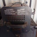 Zadní strana černého svítidla s integrovanou rukojetí. Na horní části je umístěn štítek s texty „PRECAUTIONS ESSENTIELLES“ a „SAFETY WARNINGS“. Pod n…