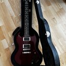 Tmavě červená elektrická kytara s tmavým hmatníkem v černém tvrdém kufru. Na hlavě nástroje je viditelné logo Gibson. Tělo kytary má dva humbuckery,…