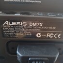 Detail zadní strany černého elektronického zařízení s nápisem ALESIS DM7X. Štítek obsahuje varovný text „CAUTION: DO NOT ALLOW THE PRODUCT TO BE EXPO…