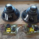 Dva černé reproduktory s kovovými rámy a magnety, na kterých jsou viditelná bílá loga. Pod nimi jsou umístěny dvě elektronické desky s osazenými souč…
