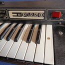Detailní pohled na klávesový nástroj s černým povrchem z texturovaného materiálu a dřevěnými bočními listy. Na horním panelu je nápis PIANO v obdélní…