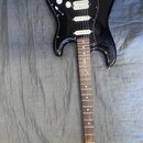 Černá elektrická kytara s černým pickguardem a hnědým hmatníkem. Na hlavě krku je vidět nápis Squier STRAT. Nástroj má tři bílé snímače, jeden přepín…