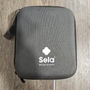 Šedý textilní obal s zipem a přivázaným popruhem. Uprostřed je bílý nápis „Sela“ a pod ním text „the soul of sound“, nad nimi je bílý symbol čtyřlíst…