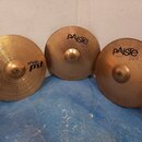 Tři mosazné činely opřené o bílou zeď na modré podlaze. Nejlevý činel vlevo nese nápisy paiste PST II a 18" CRASH RIDE. Prostřední činel má nápisy pA…