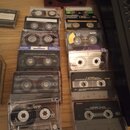 Fotografie zobrazuje skupinu audio kaset v plastových pouzdrech, rozložených na dřevěném povrchu. Jsou zde vidět značky SONY, TDK, BASF a nečitelné n…
