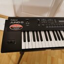 Černý digitální klavír s bílými a černými klávesami umístěný na stojanu. Na levé straně je bílý nápis Roland JUNO-D a pod ním červeno-bílá nálepka s…