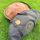 Na fotografii je kovový nástroj typu handpan v měděné barvě, umístěný v černé přepravce. Na povrchu nástroje je nápis „Handpan Design Sela“. Před nás…