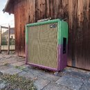 Kvadratický basový reproduktor v barevném provedení, kde je horní část zelená a spodní fialová. Na předním textilním krytu je v pravém horním rohu bí…