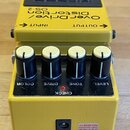Žlutý efekt pedál s černým ovládacím panelem a čtyřmi knoflíky. Na horní části je nápis „OverDrive/ Distortion“ a označení „OS-2“, v horním černém po…