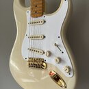 Bílá elektrická kytara typu Stratocaster s bílým pickguardem a třemi snímači s krémovými kryty. Nástroj má zlatý hardware, včetně kobylky a vložky vs…
