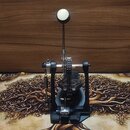Černý pedál bicích s kovovými mechanismy a bílým plstěným beaterem. Na základně pedálu je vyraženo logo, které není čitelné. Povrch nástroje vykazuje…