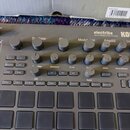 Šedý sampler/sekvencer s nápisy electribe MUSIC PRODUCTION STATION a KORG. Zařízení obsahuje několik řad šedých otočných knoflíků s popisky jako Osci…