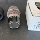 Na fotografii je mikrofonní kapsle s kovovou mřížkou, černým kroužkem a přiloženou černou pěnovou clonou. Na těle kapsle je nápis SE V7 MC2 a na krab…