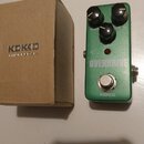 Zelený efekt pedál s nápisy OVERDRIVE a KOKKO. Na horní straně jsou tři černé ovládací knoflíky s popisky DRIVE, TONE a LEVEL. Vedle pedálu je hnědá…