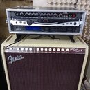 Kytarový kombozesilovač s béžovým potahem a hnědou přední síťovkou, na kterém je nápis Fender. Na horním ovládacím panelu je text Super Sonic Twin a…