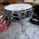 Stříbrný chromovaný snare drum s bílým blátcem značky EVANS. Na korpusu je viditelné červené logo s nápisem PREMIER. Buben je umístěn na chromovaném…
