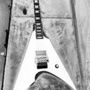 Bílá elektrická kytara s tvarem V v otevřeném kufru. Na hlavě hmatníku je viditelné logo Gibson. Nástroj má dva humbuckery, most s pružinami a jeden…