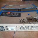Hnědý cardboardový krabic s modrou lepicí páskou. Na krabici jsou černé nápisy „FRAGILE ELECTRONIC EQUIPMENT“ a logo s textem „CREST PERFORMANCE @CRE…