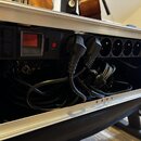 Fotografie zachycuje vnitřek hliníkového transportního kufru, ve kterém je namontována černá napájecí lišta. Lišta obsahuje červený vypínač, kontrolk…