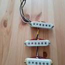 Tři elektrické snímače typu single-coil s bílými plastovými kryty a červeným základem. Každý snímač má šest viditelných kovových pólů a je připevněn…