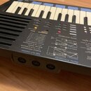 Černý digitální syntetizátor s klávesnicí, která obsahuje bílé a modré klávesy. Na ovládacím panelu je nápis DIGITAL SYNTHESIZER a označení OPERATOR…